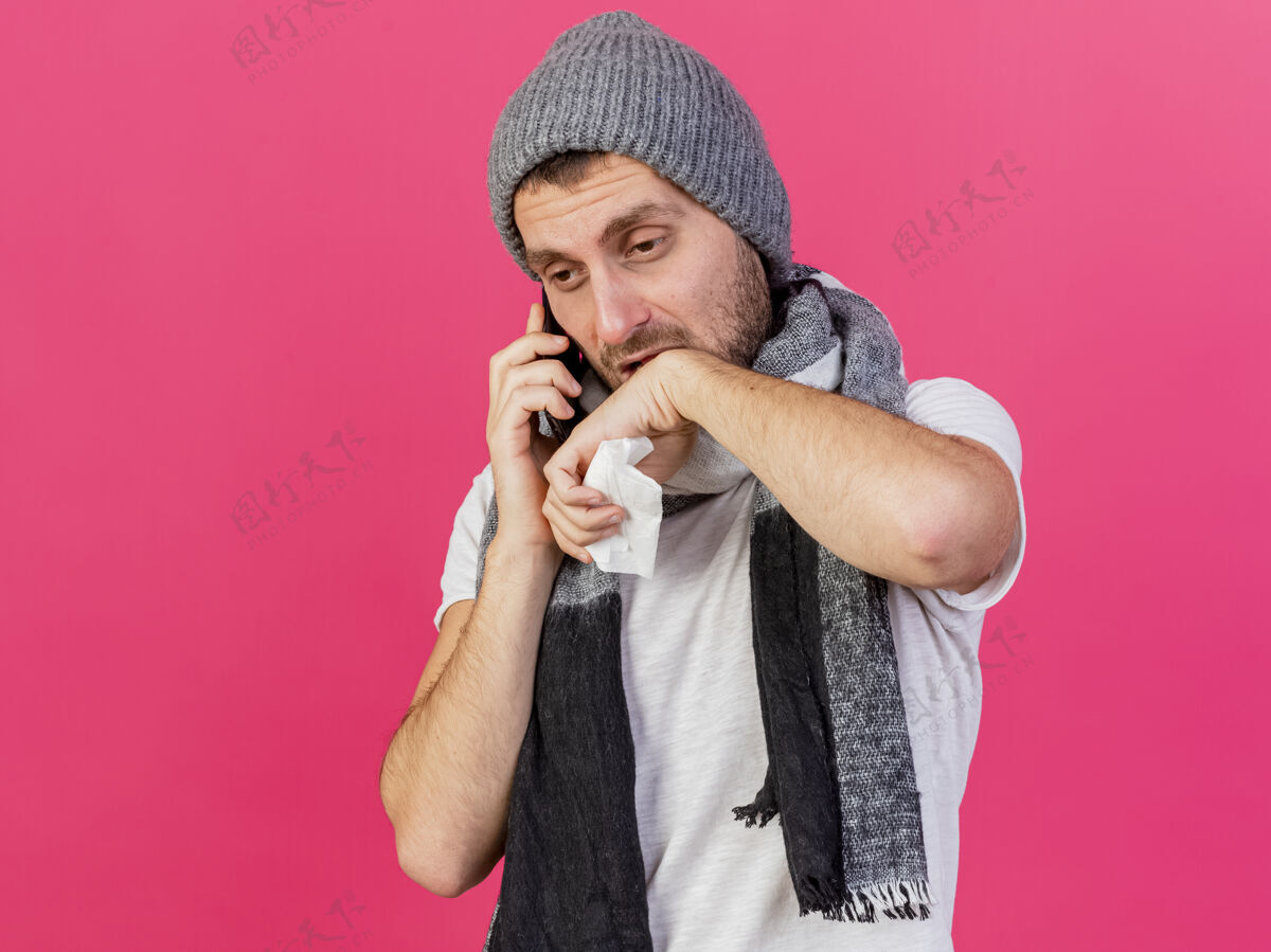 帽子戴着带围巾的冬季帽子的悲伤的年轻病夫在电话里讲话 手拿餐巾 在粉色背景上用手擦下巴穿下巴抱