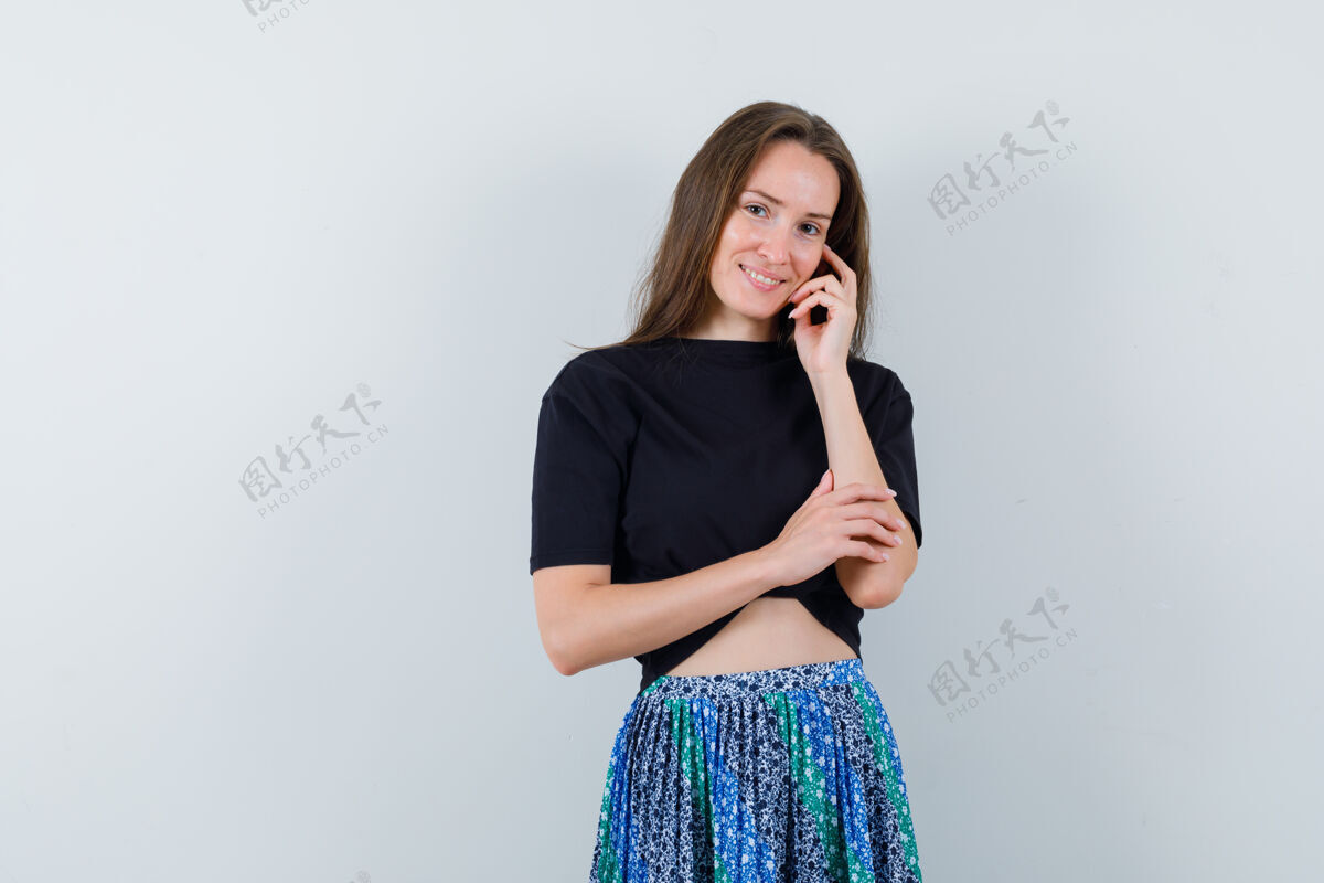 皮肤一个年轻的女人 面颊靠在手上 穿着黑色t恤和蓝色裙子在镜头前摆姿势 看起来很迷人女性手姿势