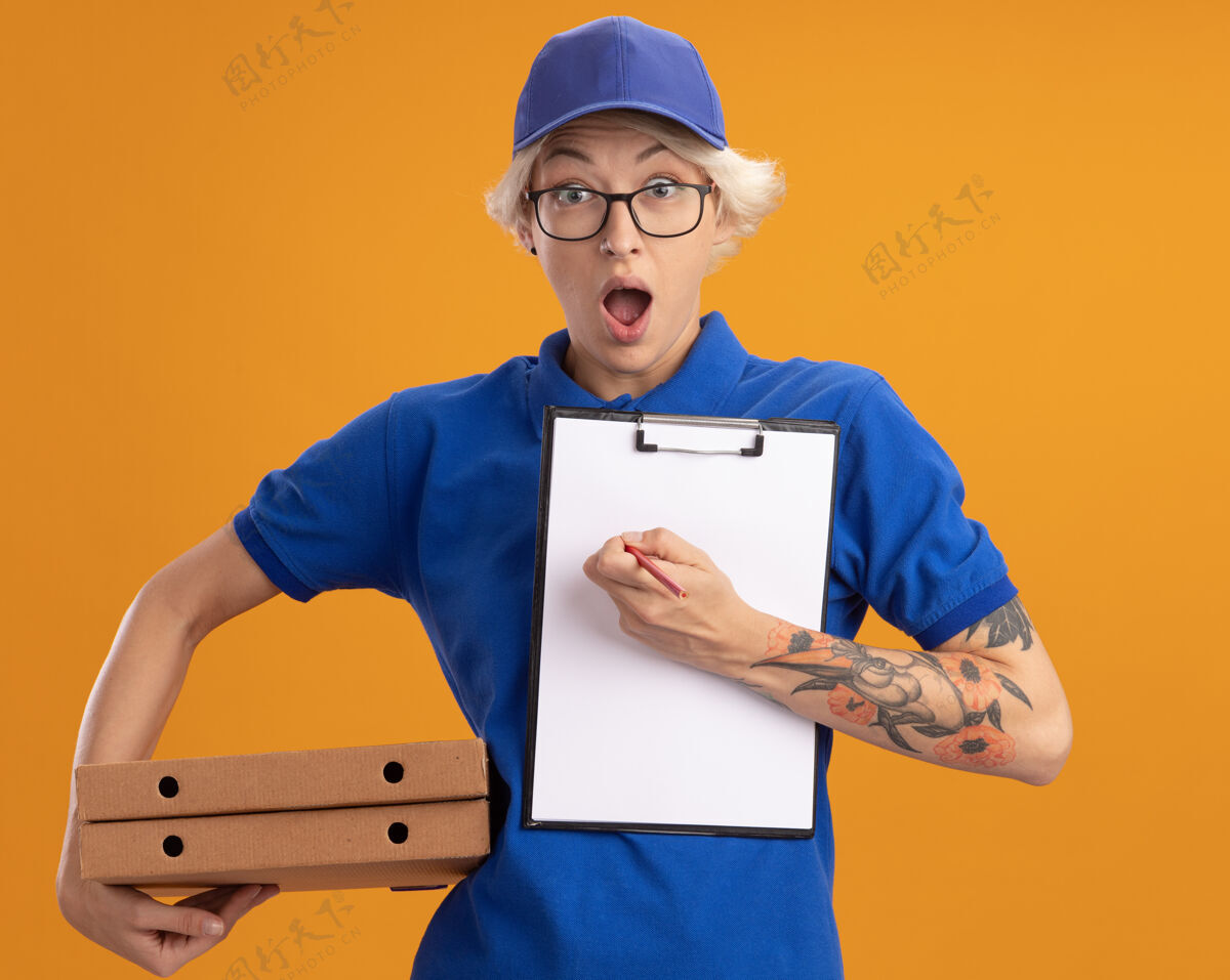 年轻身穿蓝色制服 戴着眼镜 拿着披萨盒和剪贴板 空白页和铅笔的年轻送货员 在橙色的墙上显得既惊讶又惊讶空白铅笔制服