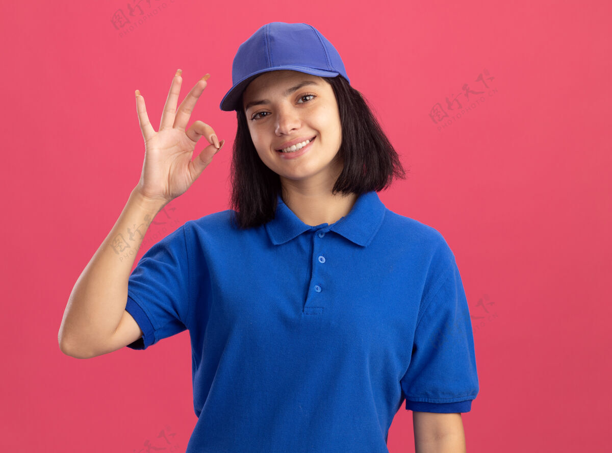 好身穿蓝色制服 头戴鸭舌帽的年轻送货员友好地微笑着 在粉红色的墙上立着ok标志朋友女孩递送