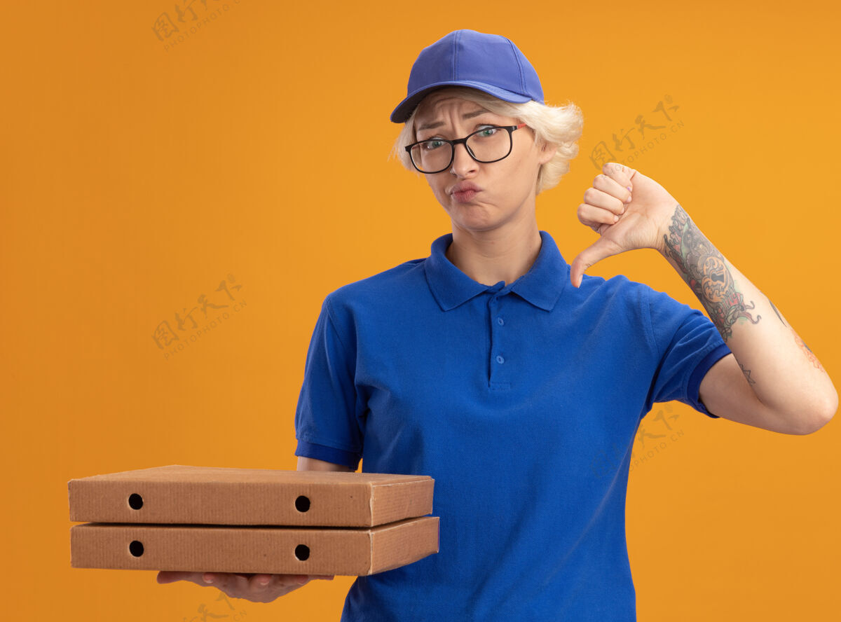 女人身穿蓝色制服 戴着眼镜 拿着比萨饼盒的年轻送货员不高兴地把大拇指放在橙色的墙上制服不高兴放下