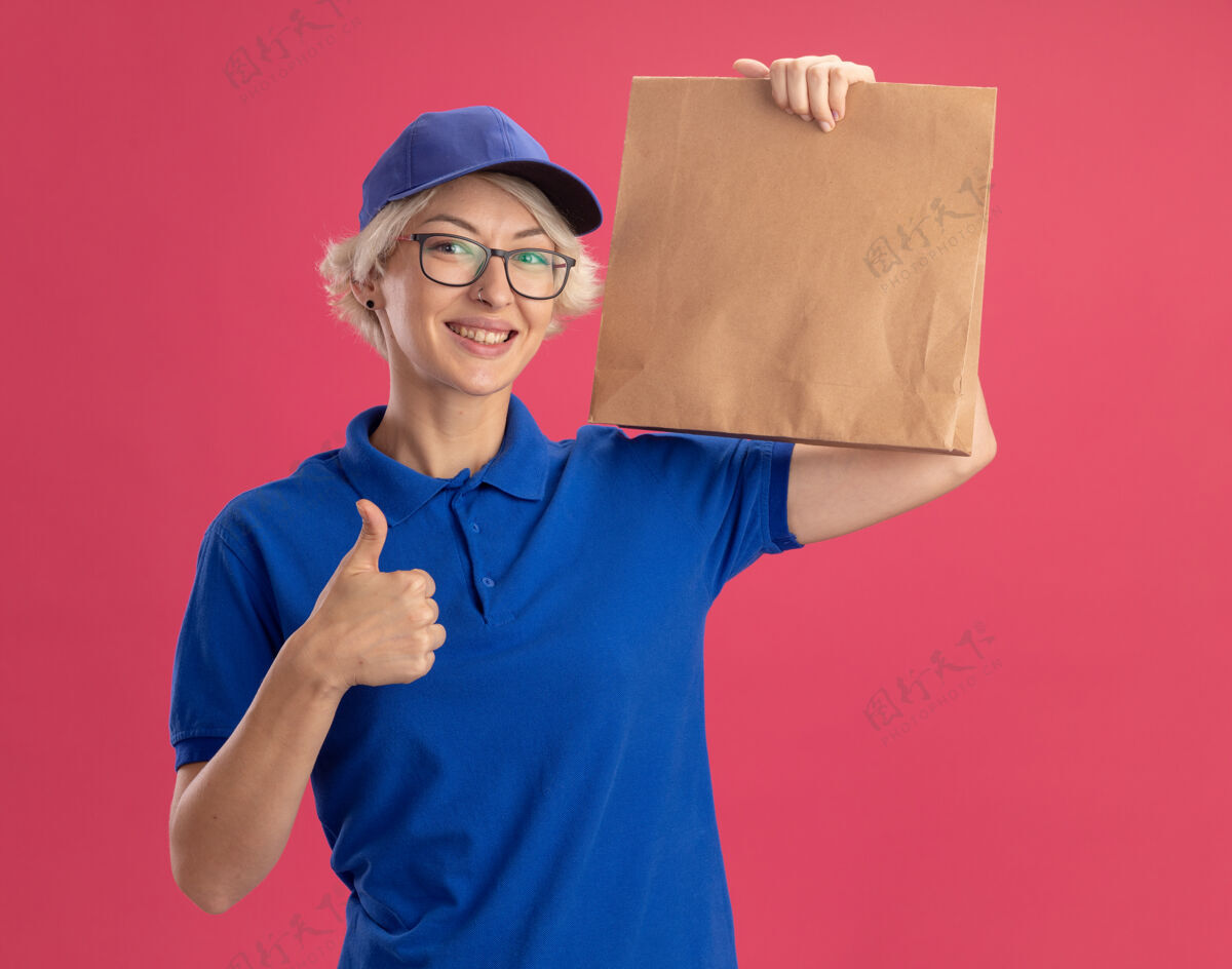 帽子身穿蓝色制服 戴着眼镜 拿着纸包 兴高采烈地向粉色墙壁竖起大拇指的年轻送货员包装微笑送货