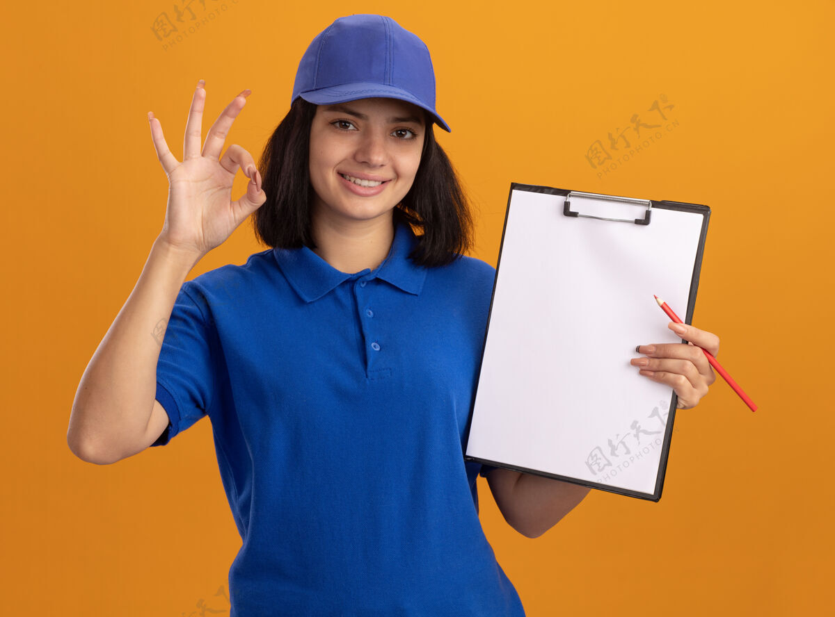 年轻身着蓝色制服 戴着帽子的年轻送货女孩拿着空白页和铅笔的剪贴板微笑着 橙色的墙上挂着“ok”的牌子女孩空白铅笔