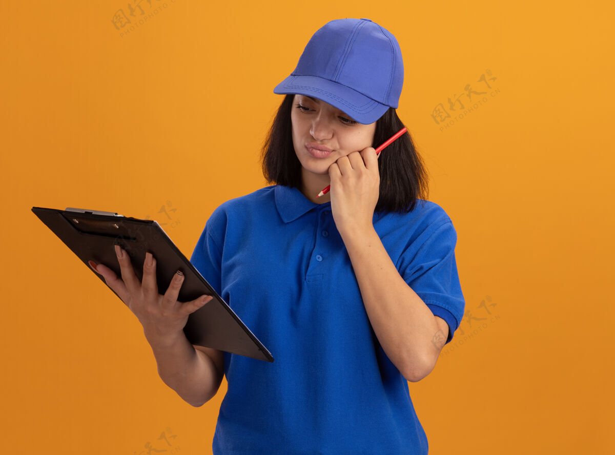 制服身穿蓝色制服 戴着帽子 拿着剪贴板和铅笔的年轻送货女孩站在橙色的墙上 看起来很困惑剪贴板年轻铅笔