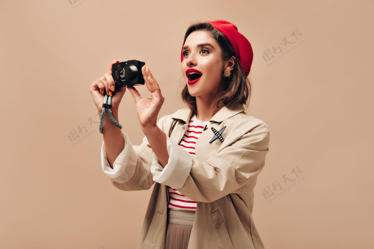 休闲一位身着盛装的女士在孤立的背景上拿着相机身着红色明亮贝雷帽的漂亮女孩在米色背景上拍照米色背景贝雷帽相机