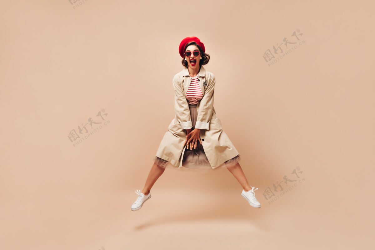 米色背景一个戴着贝雷帽 穿着外套 在米色背景下蹦蹦跳跳的情绪化女人一个戴着墨镜 穿着白色运动鞋 留着深色头发的聪明女孩摆姿势太阳镜休闲贝雷帽