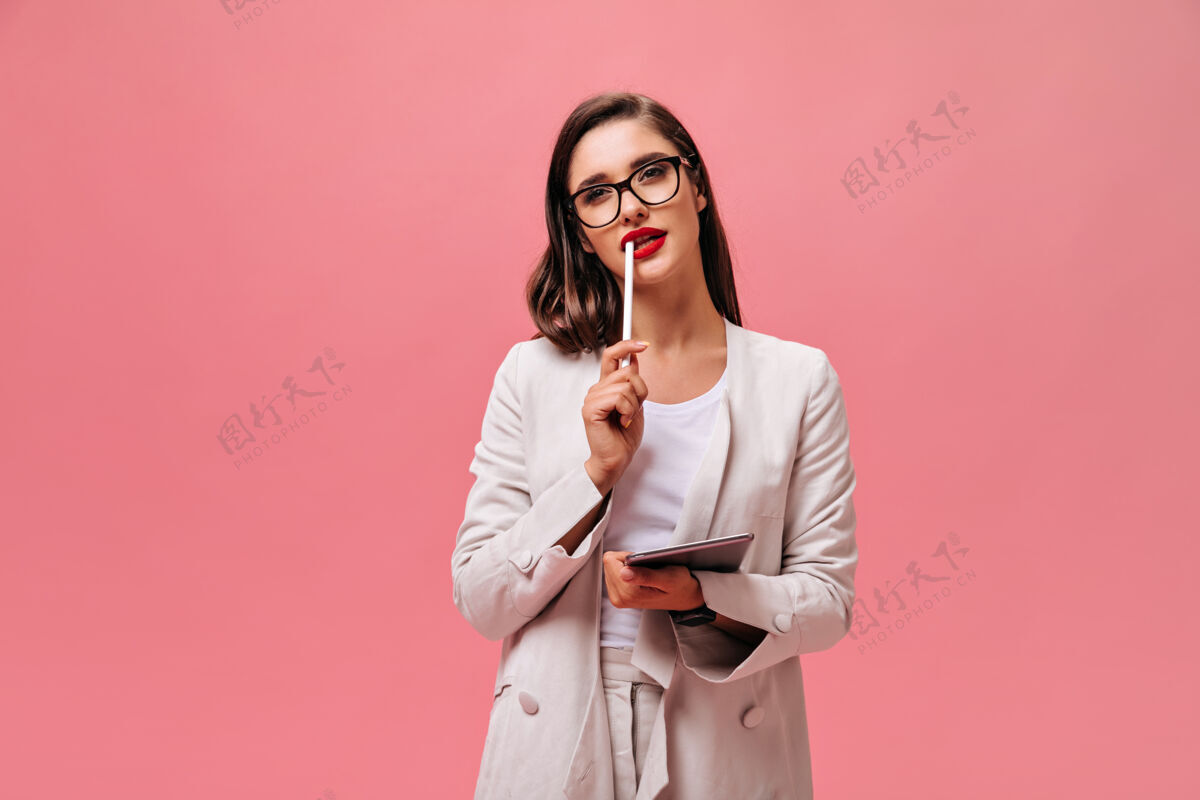 时尚迷人的年轻女子 深色头发 穿着经典西装 戴着眼镜 手持电脑平板电脑 背景是孤立的粉色粉色站立平板电脑