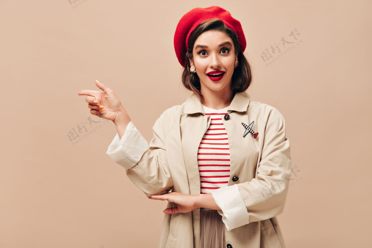 贝雷帽棕色眼睛的女士戴着红色贝雷帽 穿着米色战壕 指着地方发短信穿着条纹衣服的聪明年轻女士摆姿势拍照女孩卷发女性