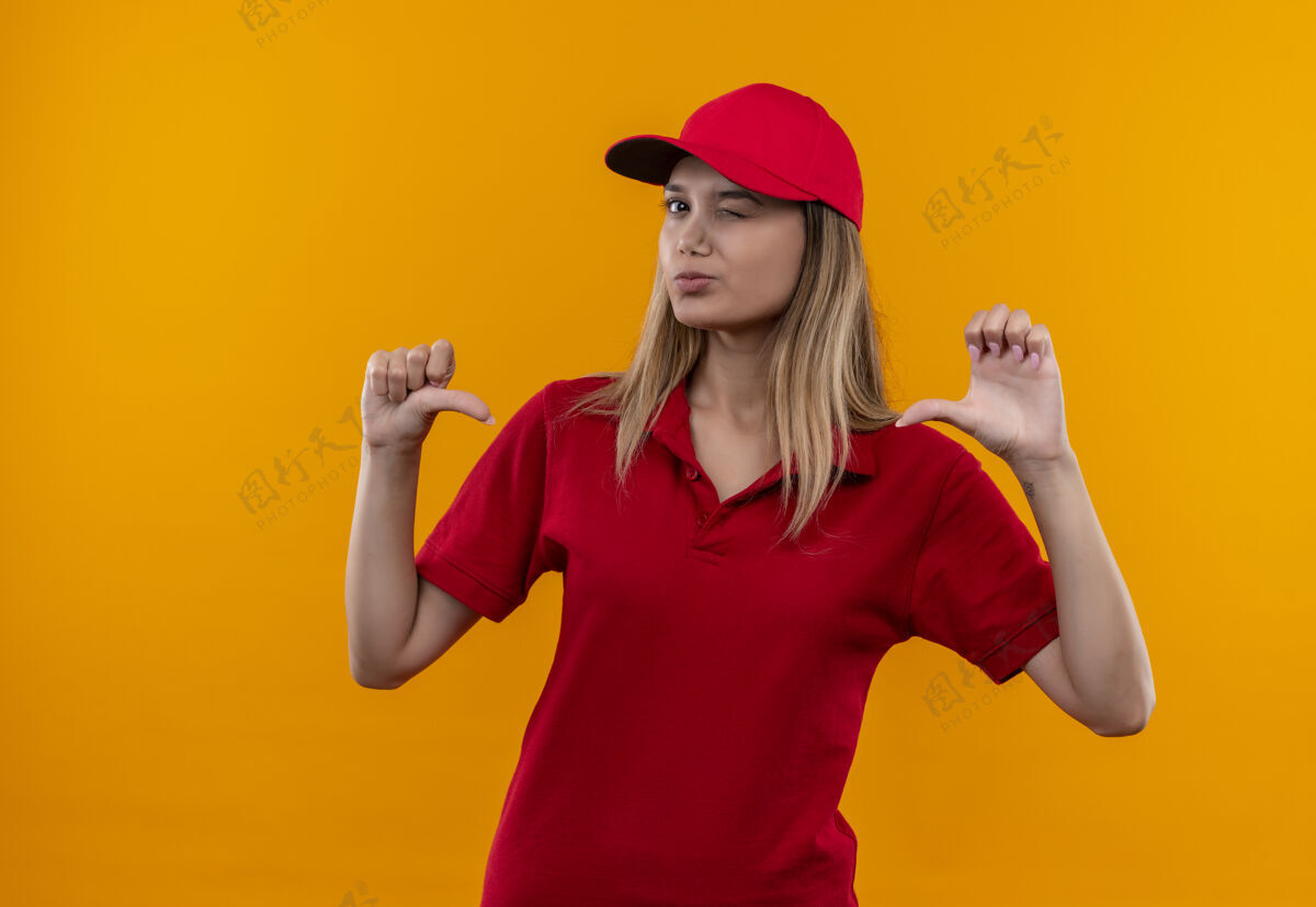 穿戴着红色制服和帽子的年轻送货女孩眨着眼睛 把自己孤立在橙色的背景上年轻女孩眨眼