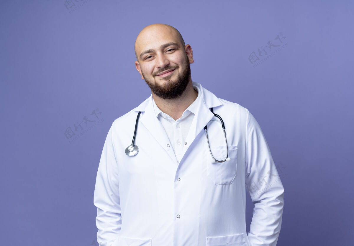 医疗高兴的年轻秃头男医生穿着医用长袍和听诊器隔离在蓝色背景上男性秃头请戴上