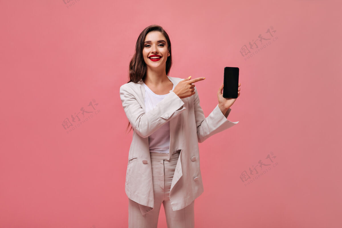 女士身着米色西装的迷人女士在粉色背景上展示智能手机在手机上展示了一位化妆亮丽的时尚女士电话卷发年轻