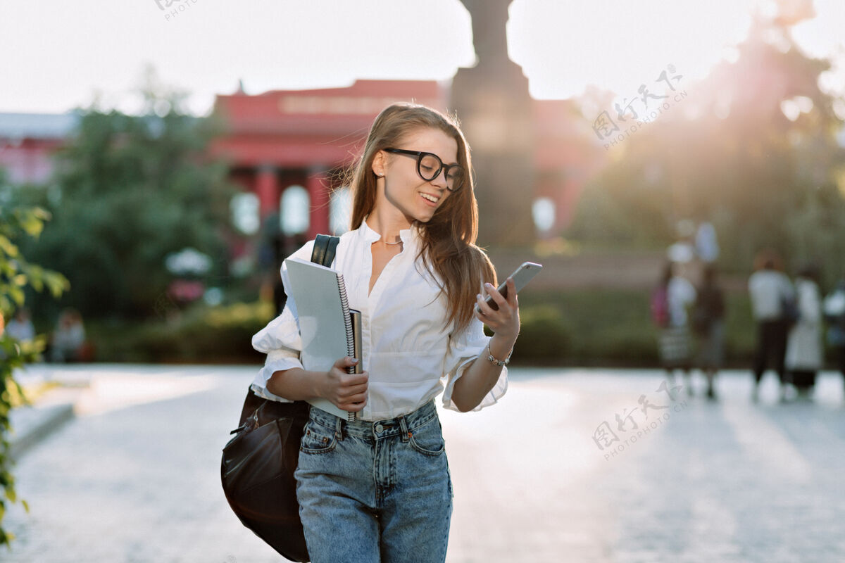 保持带着眼镜和衬衫的棕发微笑的学生带着笔记本电脑和背包在户外散步 在阳光下看着智能手机Campus大学公园
