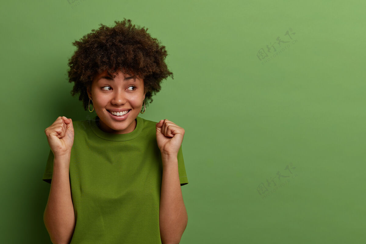 成功开朗的黑皮肤女人 非洲式发型 握紧拳头 等待积极的结果 高兴地看着一边 穿着休闲服 隔离在绿色墙上 为您的广告复制空间表情情绪面部