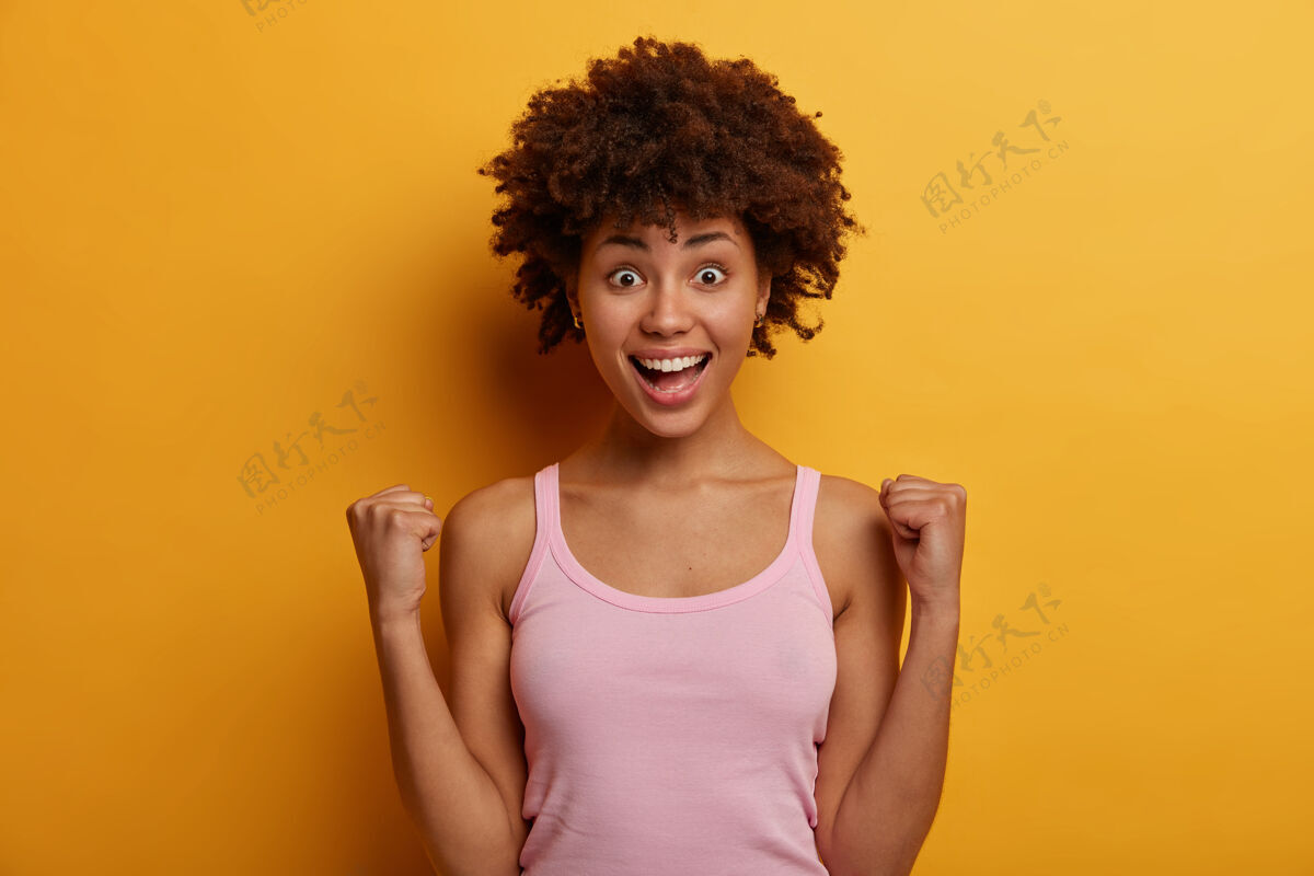 微笑一个快乐的黑皮肤非裔美国女人胜利地握紧拳头 高兴地成为冠军并赢得第一名 高兴地微笑着 为积极的消息欢呼 穿着休闲背心女人正面种族