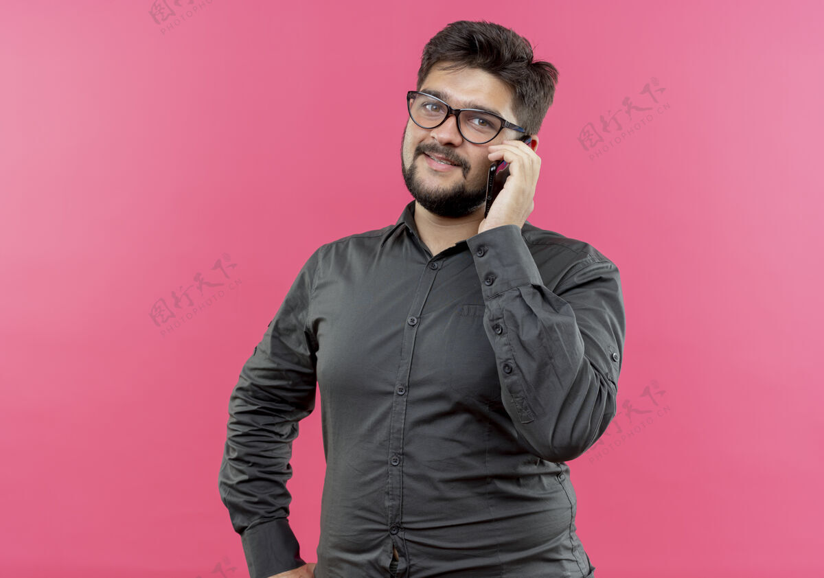 臀部戴着眼镜的年轻商人在电话里讲话 手放在屁股上 粉红色的背景上孤立着电话手说话