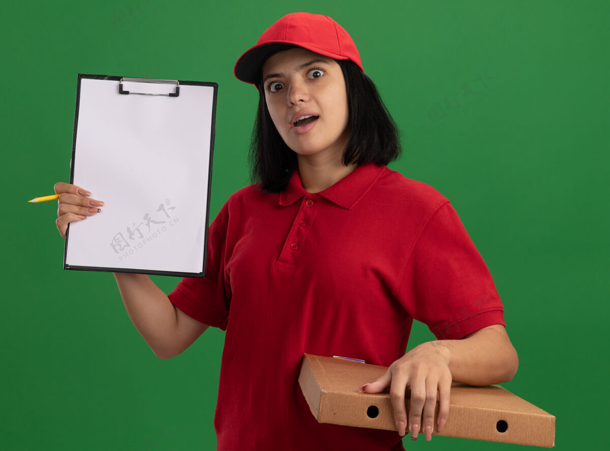 制服穿着红色制服 戴着帽子 拿着披萨盒 拿着铅笔的小女孩惊讶地站在绿色的墙上立场显示举行