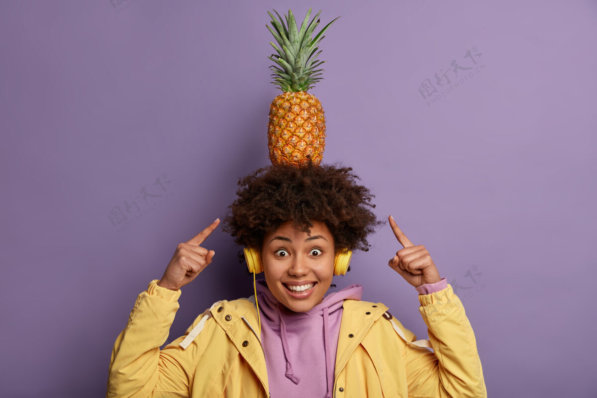 周末看我怎么能正面开朗的十几岁的美国黑人女人指着头上的菠萝 戴着耳机听音频 穿上连帽衫和夹克 隔着紫色的墙壁 一个人玩旋律头运动衫