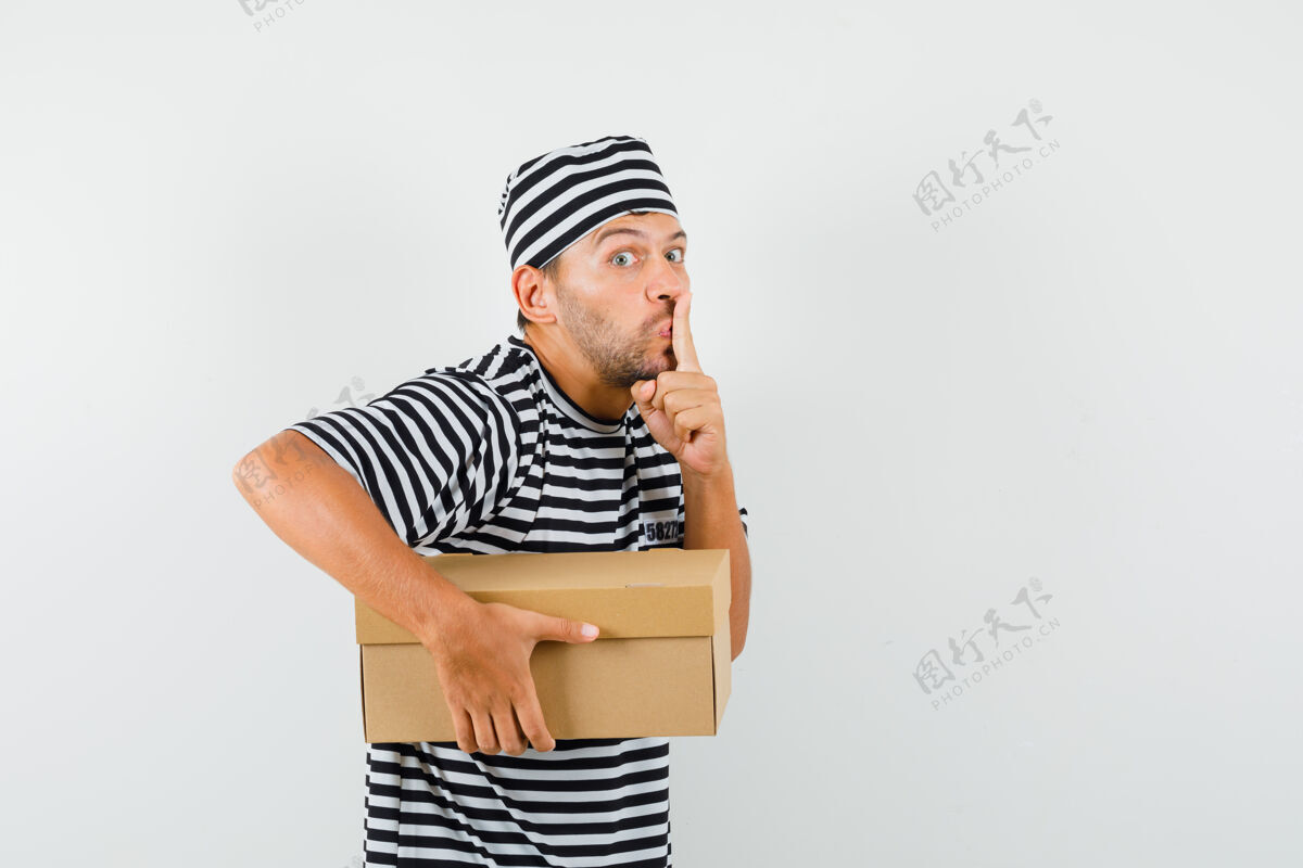 帽子一个年轻人拿着一个纸板箱 戴着条纹t恤帽子 表现出沉默的姿态 看上去很小心成功盒子现代