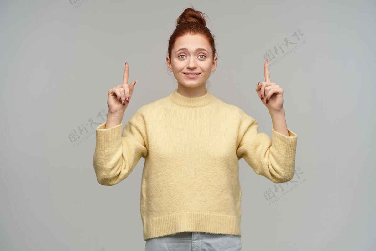 成人一幅迷人的成年红发女孩的肖像 头发盘成一个小髻 穿着淡黄色的毛衣和牛仔裤指着复制空间 隔着灰墙微笑快乐年轻
