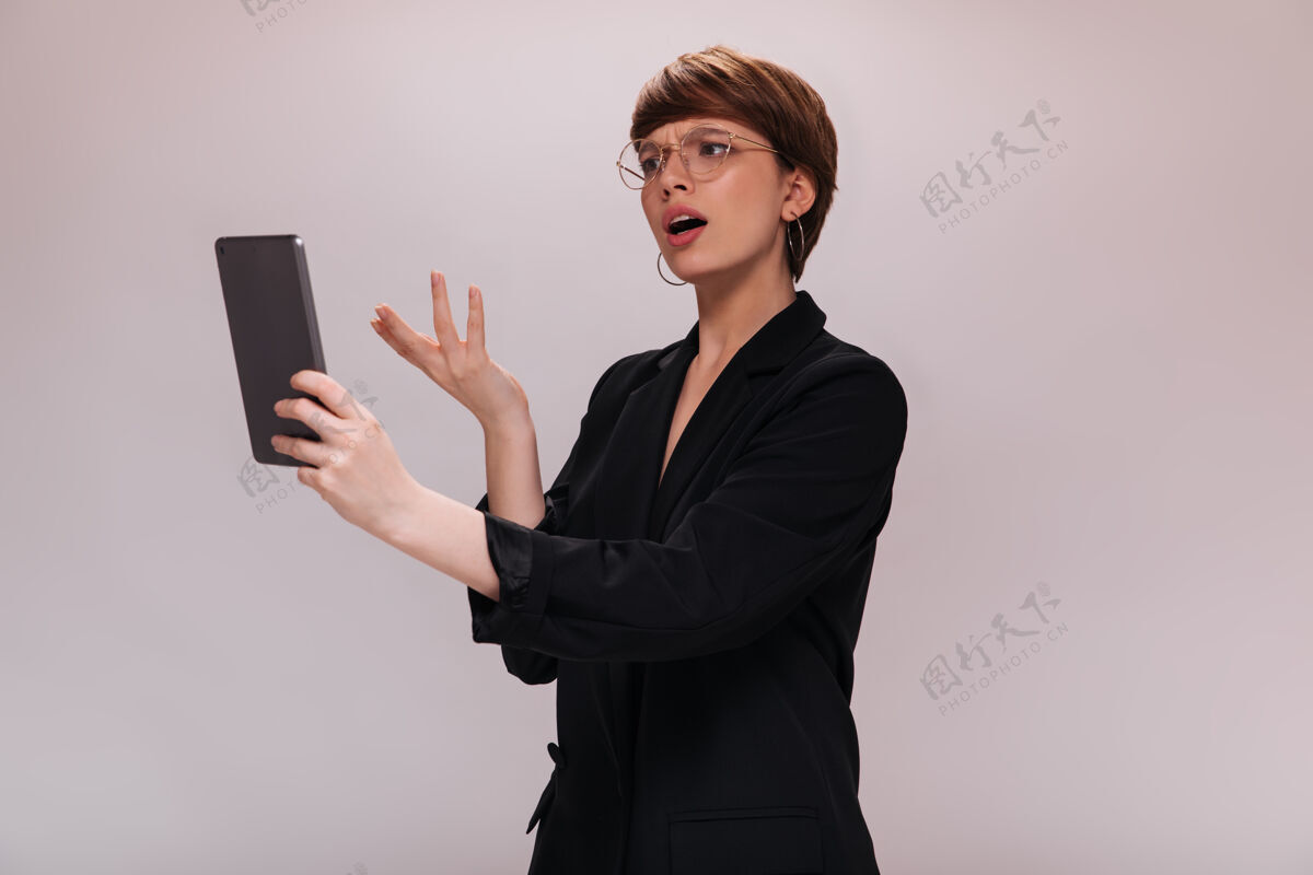 优雅女士误会地看着平板电脑屏幕穿着黑色夹克的商务女士在孤立的白色背景上摆姿势套装白色站立