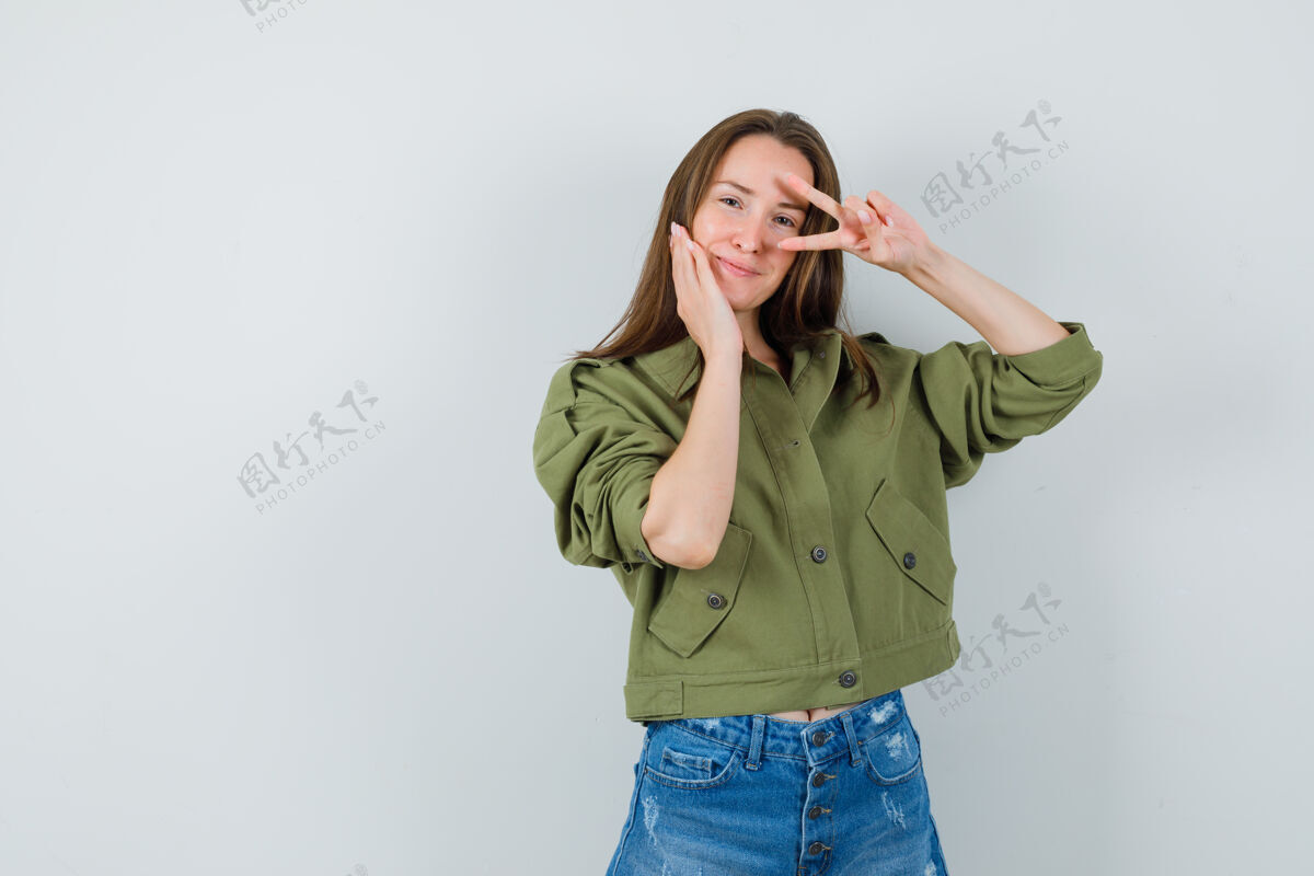 近穿着绿色夹克短裤的年轻女士在眼睛附近展示v型标志 看起来很自信人性化连衣裙在一起