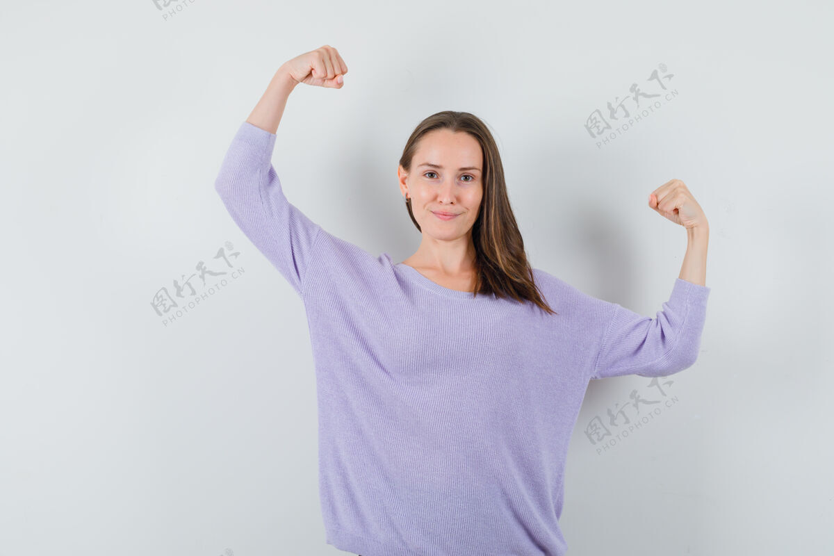 而年轻的女性举起她的手臂 同时显示在淡紫色衬衫手臂肌肉 看起来很强大前视图姿势自信肌肉