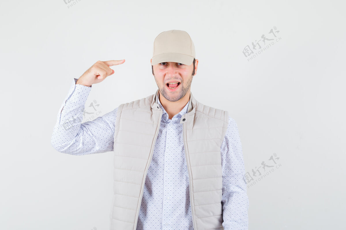 年轻年轻人指着他的帽子 在衬衫 无袖夹克 帽子和乐观的样子 前面的看法胡须模特休闲