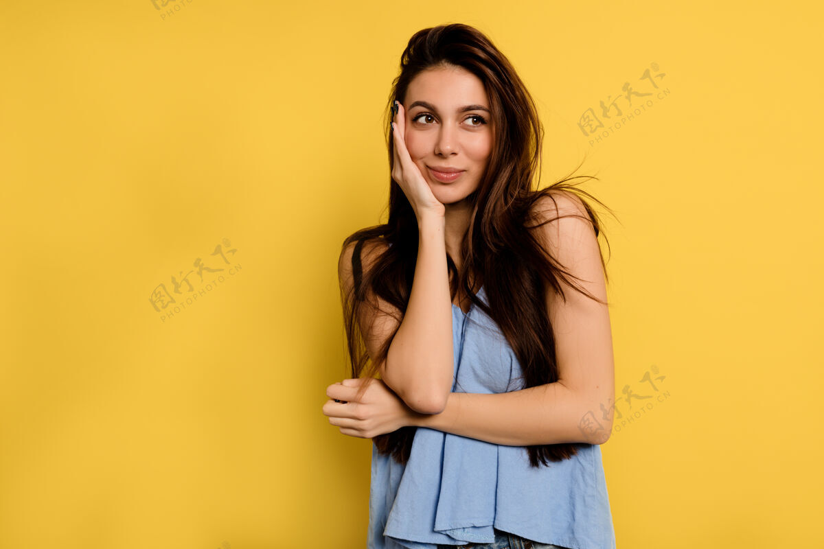 快乐穿着蓝色衬衫的美丽快乐的女人用手捂着脸 梦幻般地看着黄色墙上的镜框成功模特手臂