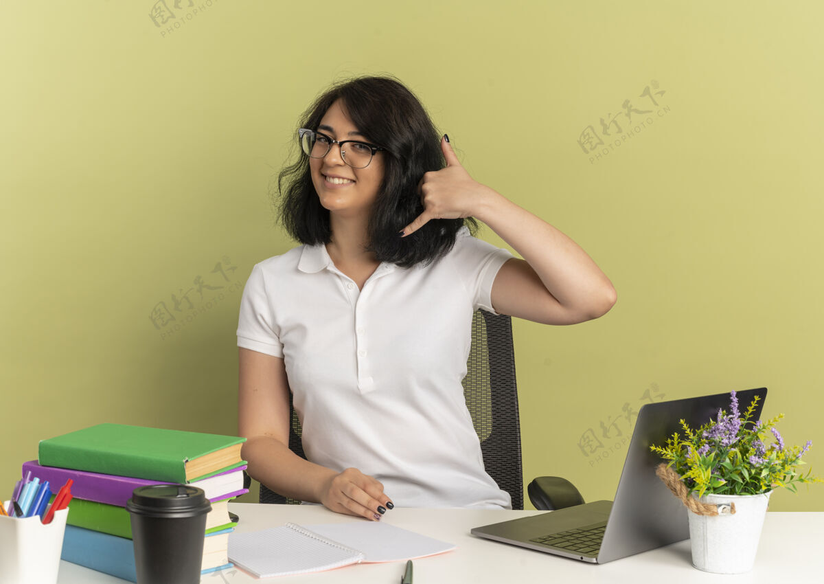 眼镜年轻的高加索女孩戴着眼镜 面带微笑 坐在书桌旁 拿着学习工具 手势 手机 手势 绿色 还有复印空间坐女学生手