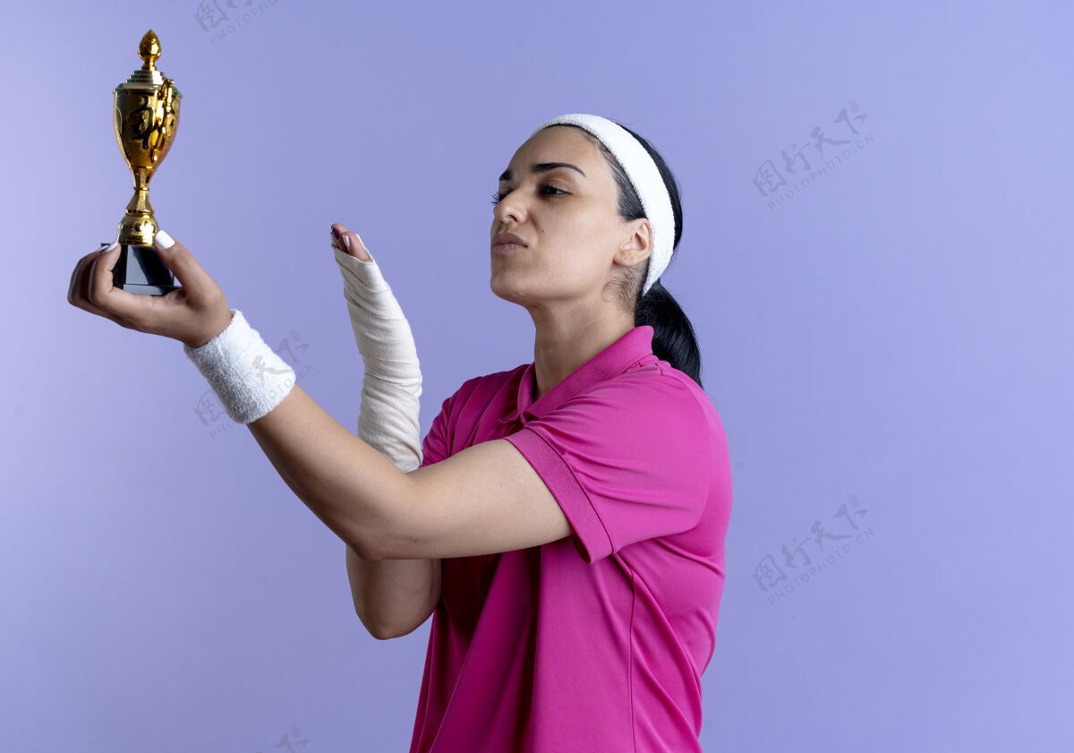 运动年轻自信的高加索运动女性戴着头带和腕带拿着并指着紫色的冠军杯穿女子腕带