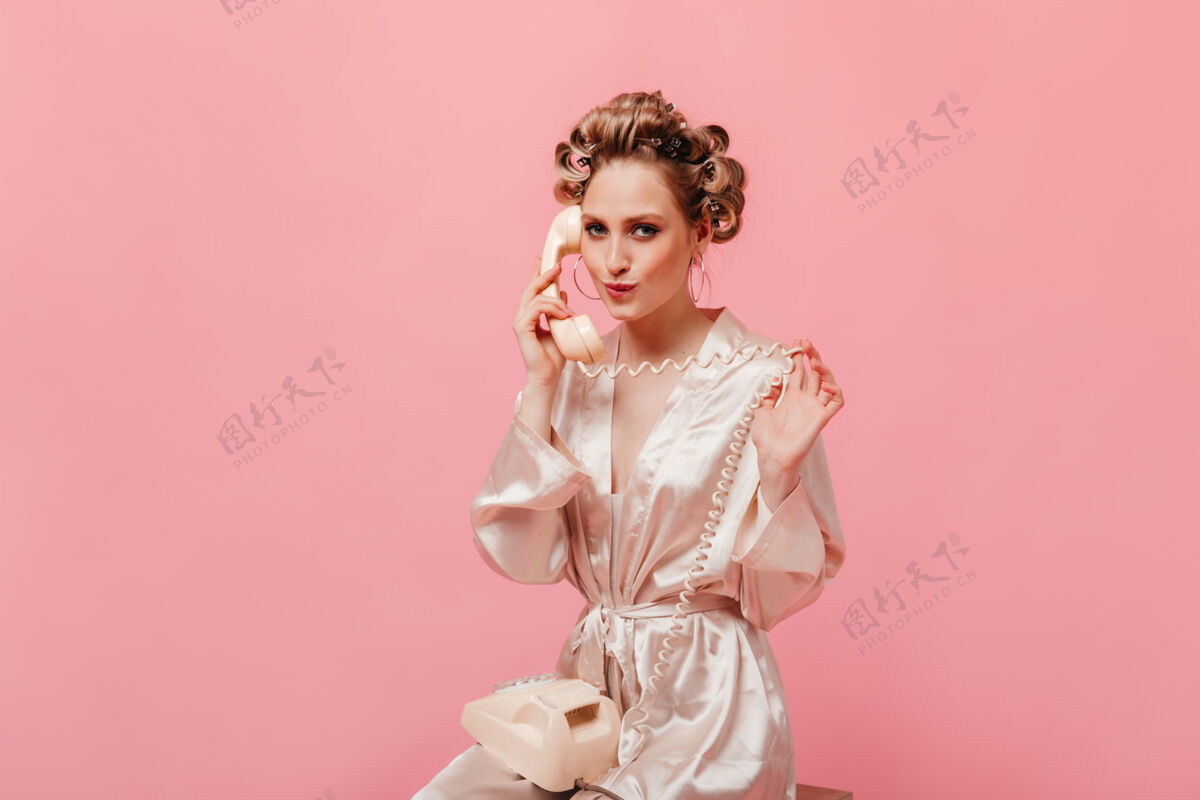 时尚穿着丝绸睡衣的美女在粉红色的墙上撒娇地讲电话移动粉色浴袍