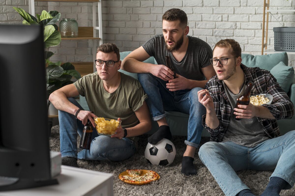 足球三位男性朋友一边吃零食和啤酒 一边看电视上的体育节目看啤酒男
