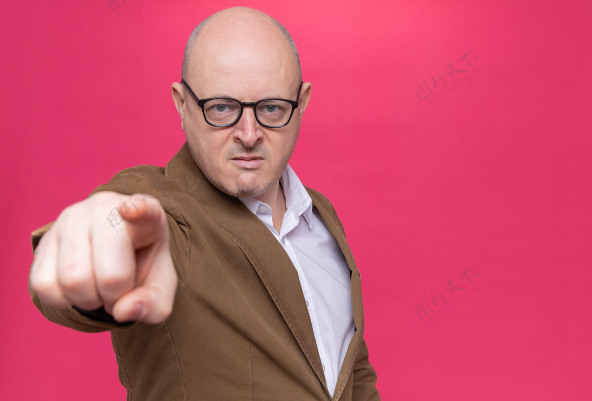 眼镜愤怒的中年光头男子穿着西装戴着眼镜 食指指着前面站在粉红色的墙上愤怒手指站着
