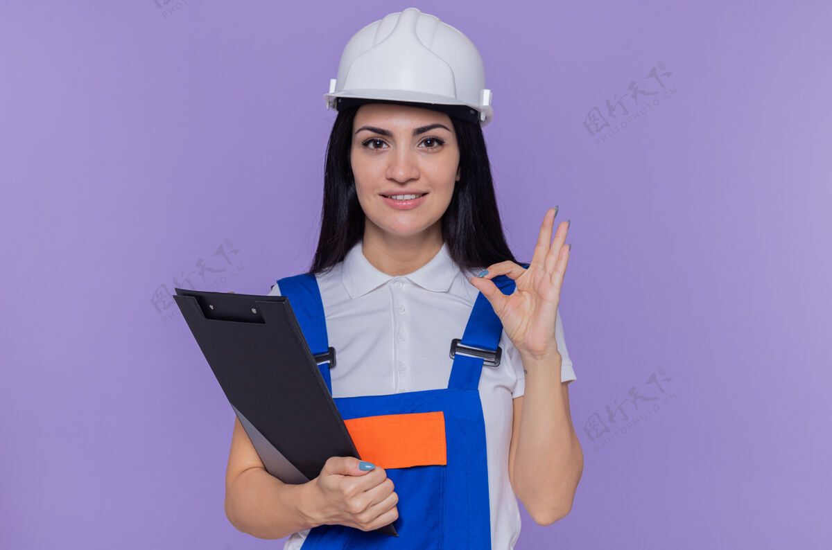 建筑工身穿施工制服 头戴安全帽 拿着写字板和铅笔的年轻建筑工人看着前方 脸上洋溢着幸福和积极的微笑 自信地站在紫色的墙上做着“ok”的手势Ok铅笔积极