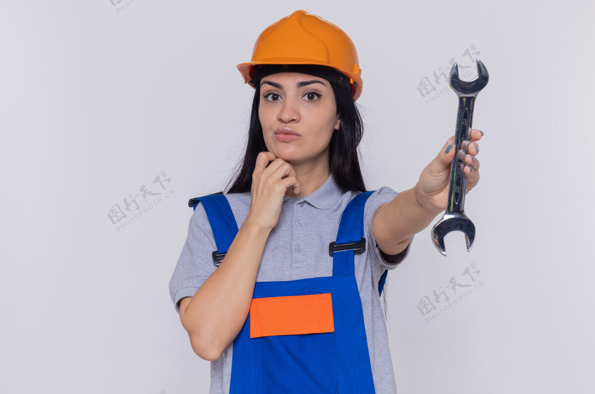 扳手年轻的建筑工人妇女穿着建筑制服 戴着安全帽 拿着扳手 站在白色的墙上 困惑地看着前面建筑工站立建筑