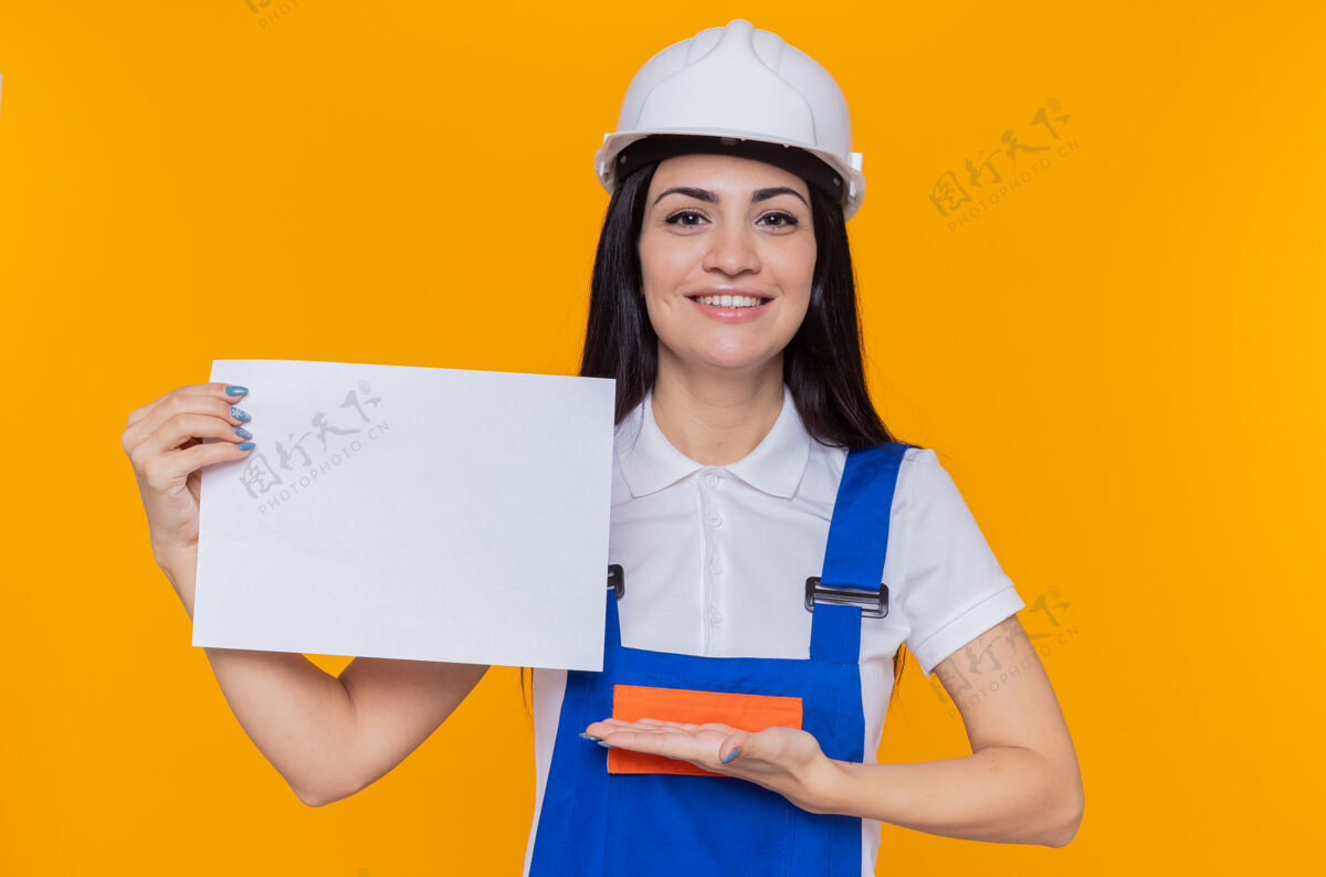 空白身穿施工制服 头戴安全帽的年轻建筑工人手举空白页 微笑着自信地站在橙色的墙上建筑工手臂信心