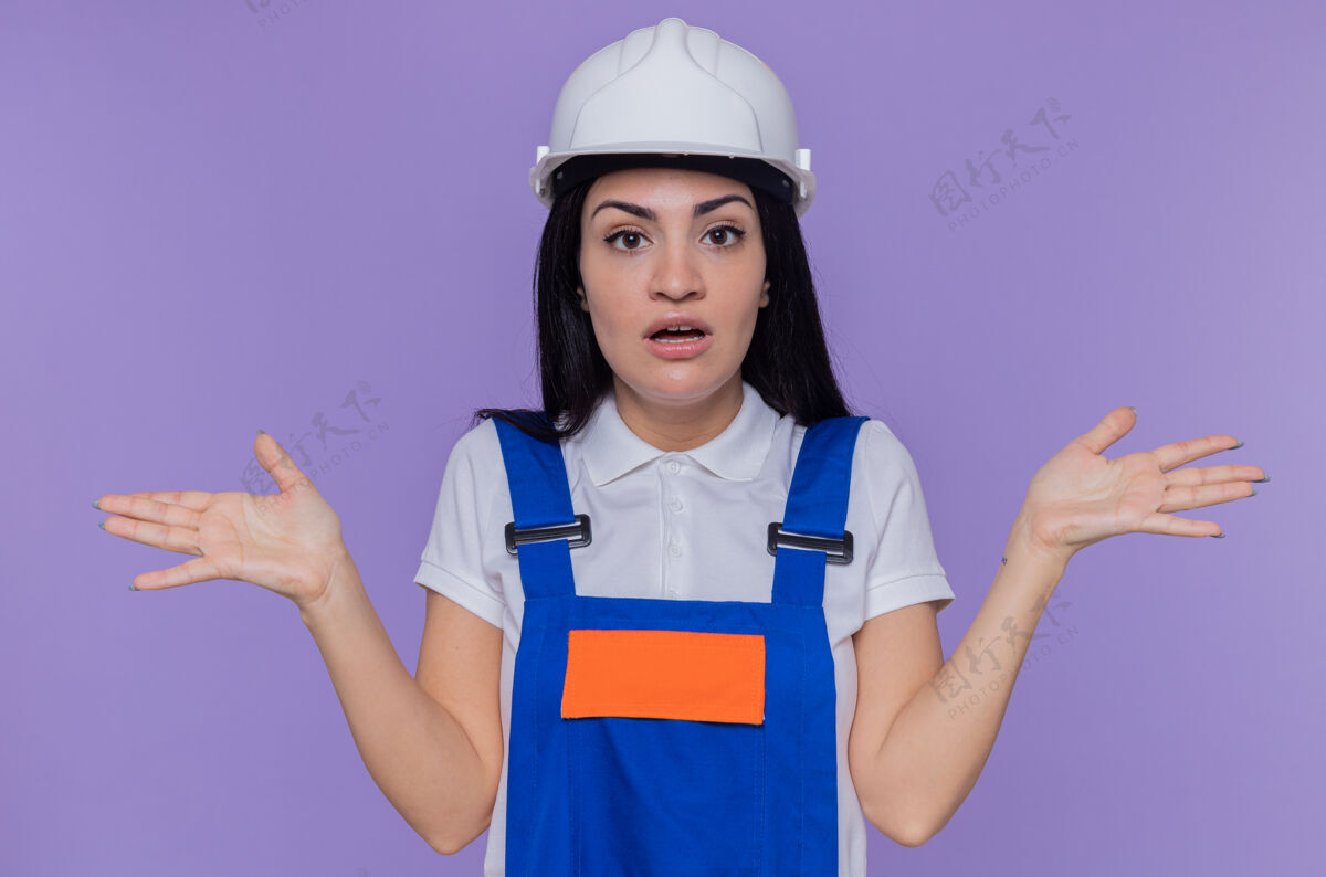 手臂身穿施工制服 头戴安全帽的年轻建筑女工人站在紫色的墙上 困惑地看着前方 举起双臂困惑年轻举起