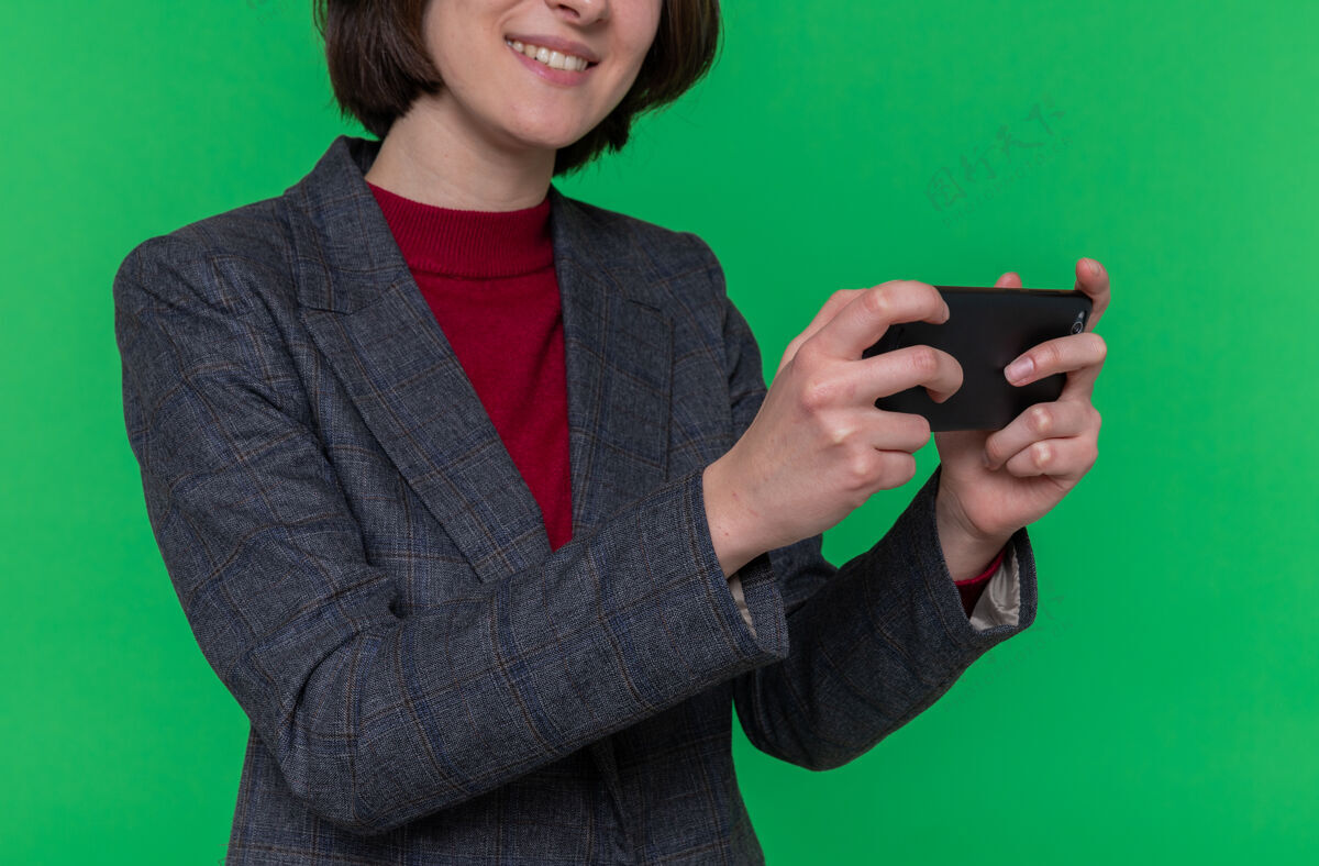 女人一个留着短发的年轻女人穿着灰色夹克玩着智能手机游戏快乐地站在绿色的墙上游戏快乐短裤