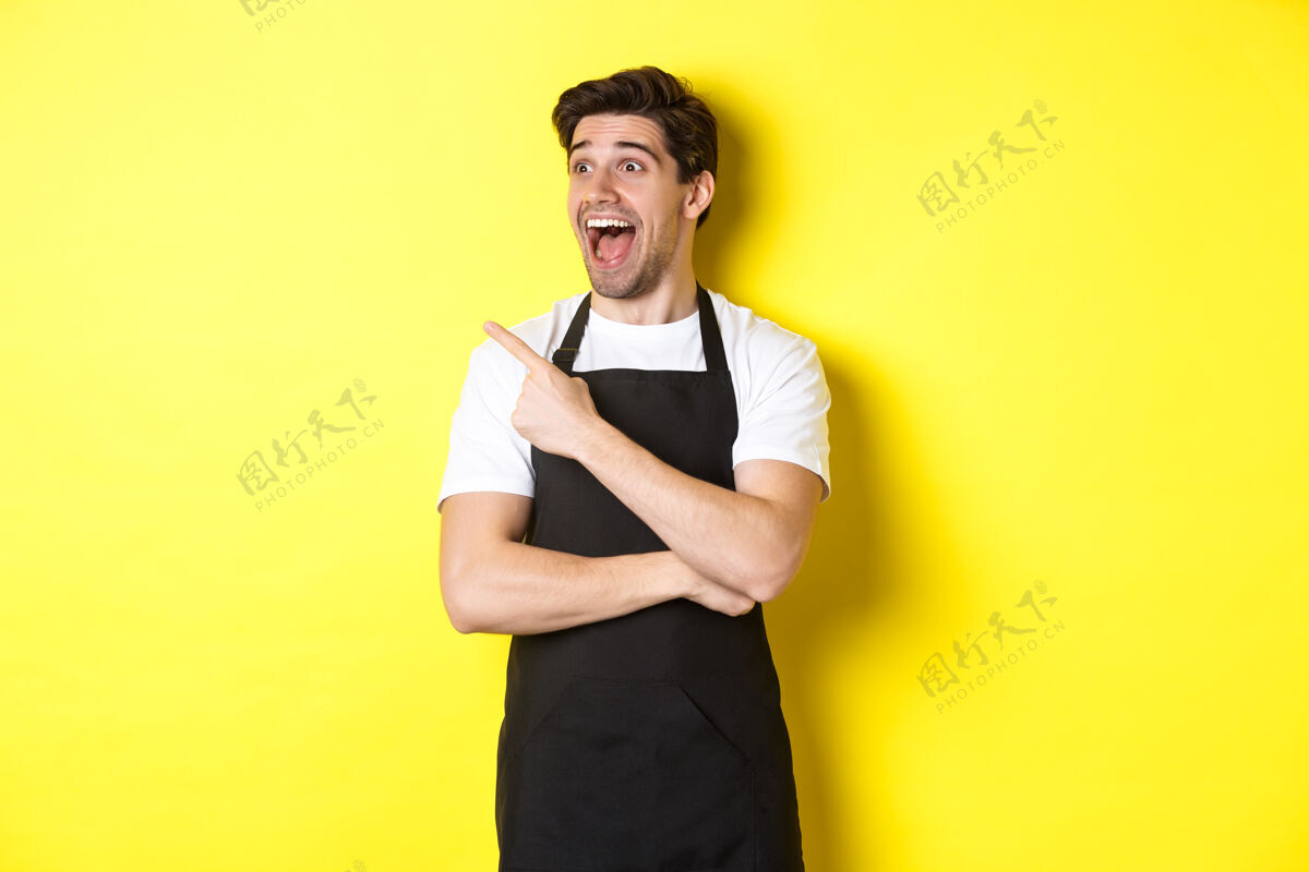 厨师穿着黑色围裙的服务员正在查看促销活动 手指着你们的商标 站在黄色背景上看左边成人男人工作室