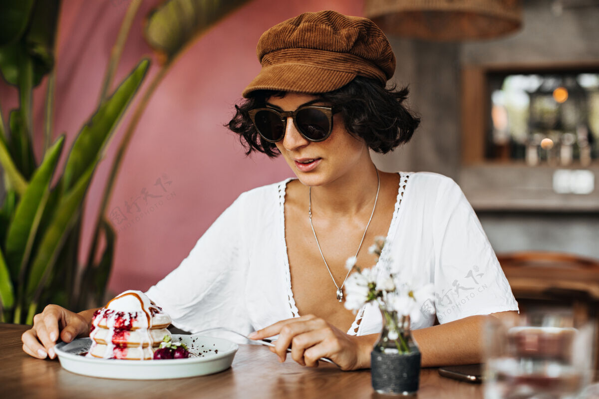 浆果迷人的深色皮肤 戴着墨镜和帽子的棕褐色女人在咖啡馆吃甜点奶油盘子美味