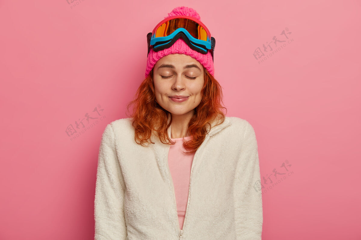 室内迷人的红发女人戴着暖和的帽子 滑雪镜和白色套头衫跳伞者请表达