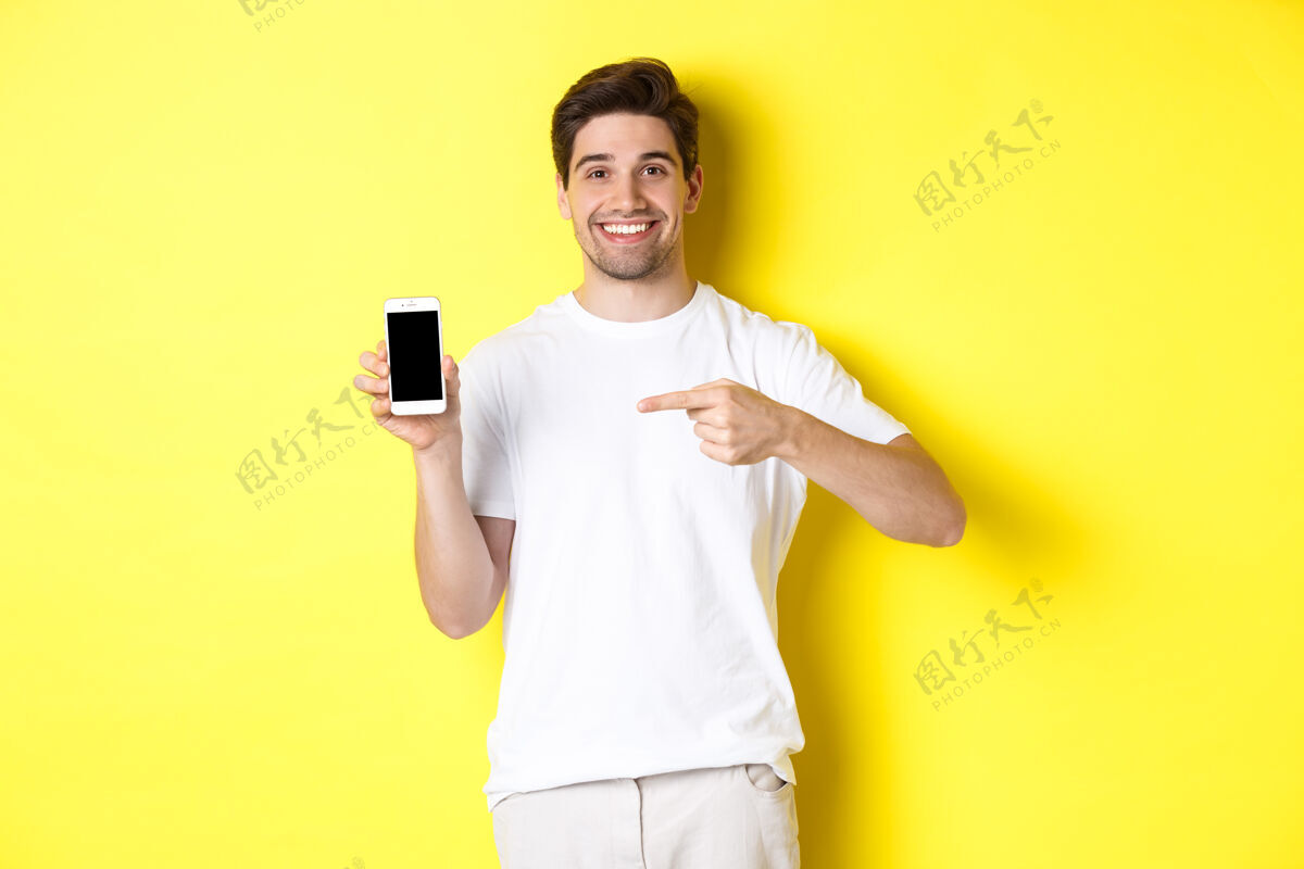 时尚一个漂亮的年轻人用手指着智能手机屏幕 展示一个应用程序 站在对面黄色模特自信