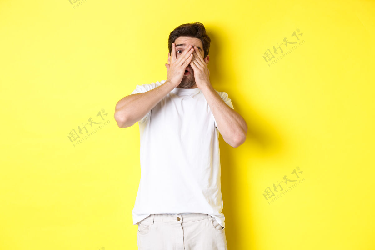 男人尴尬的家伙闭上眼睛 但透过手指偷看一些尴尬的东西 站在黄色背景上男人男人成年人