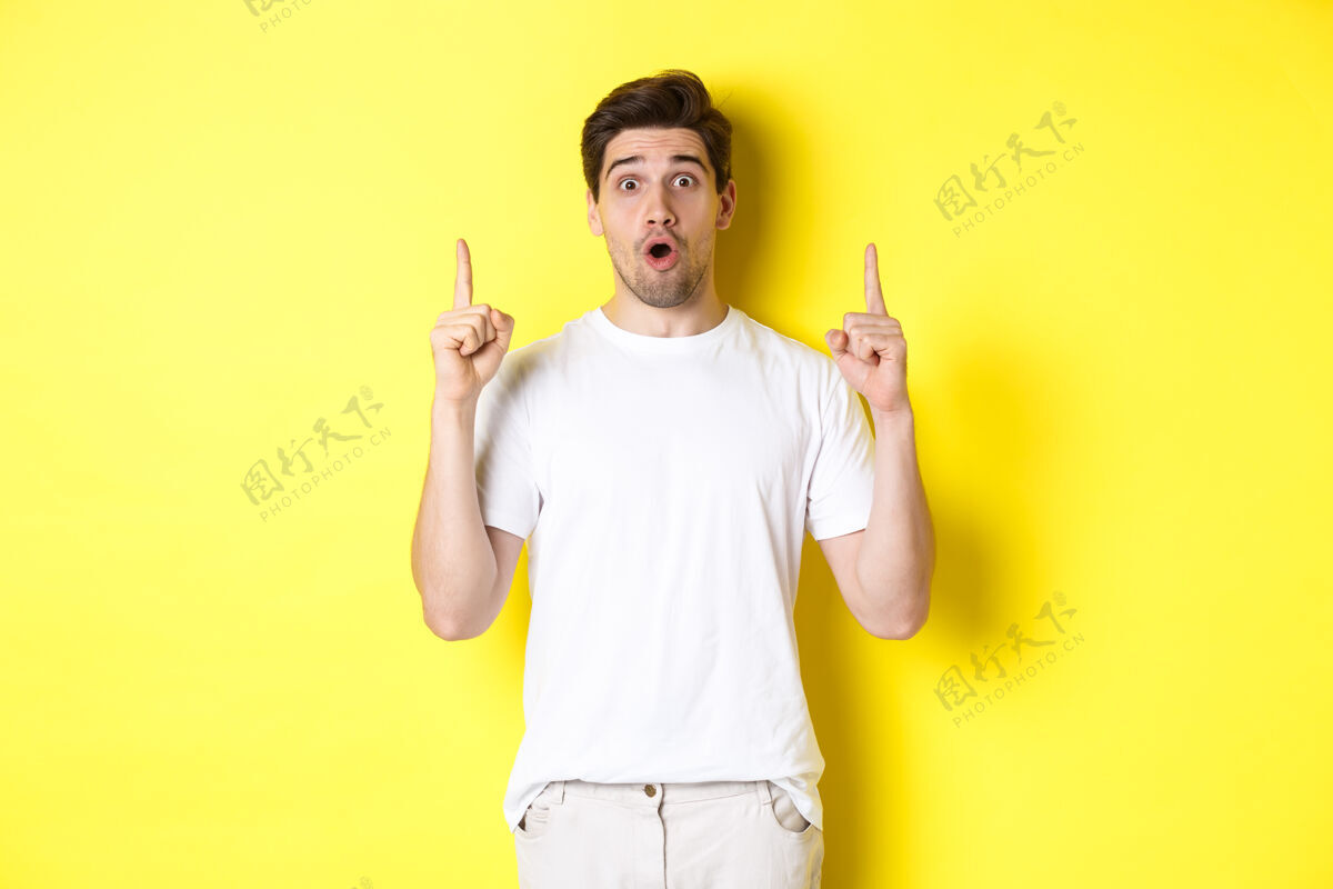 男人穿着白色t恤的帅哥 手指向上 对广告感兴趣 站在黄色背景下男人惊喜时尚