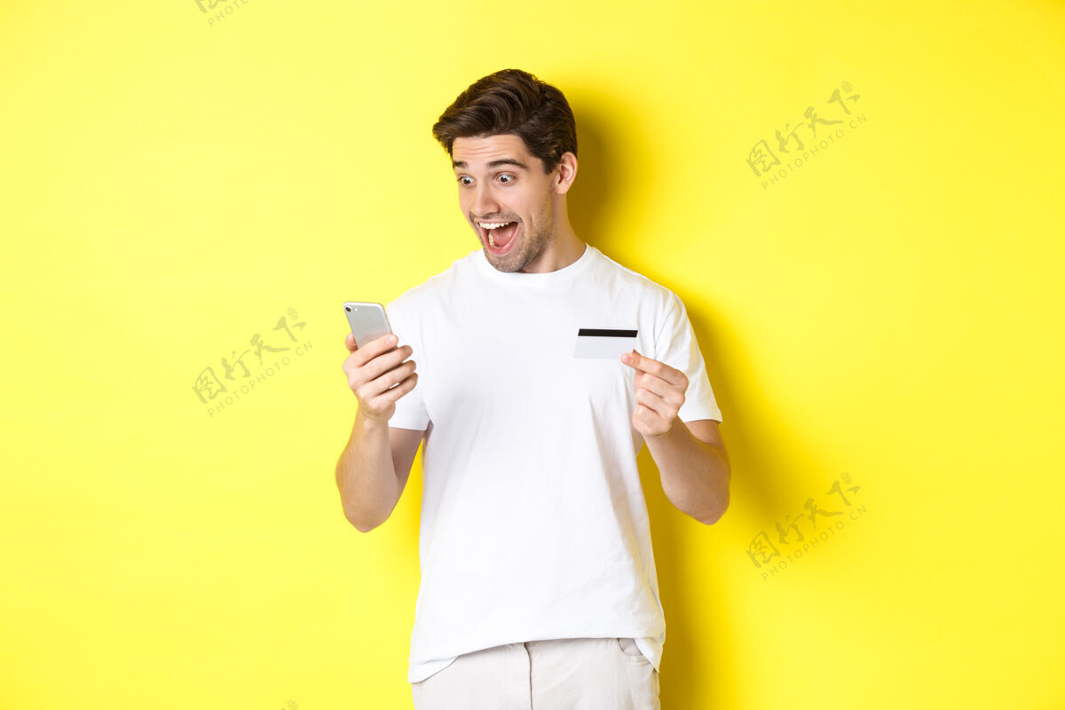 年轻男人惊讶地看着智能手机 网上购物 拿着信用卡 站在黄色背景上复制空间表情工作室站着