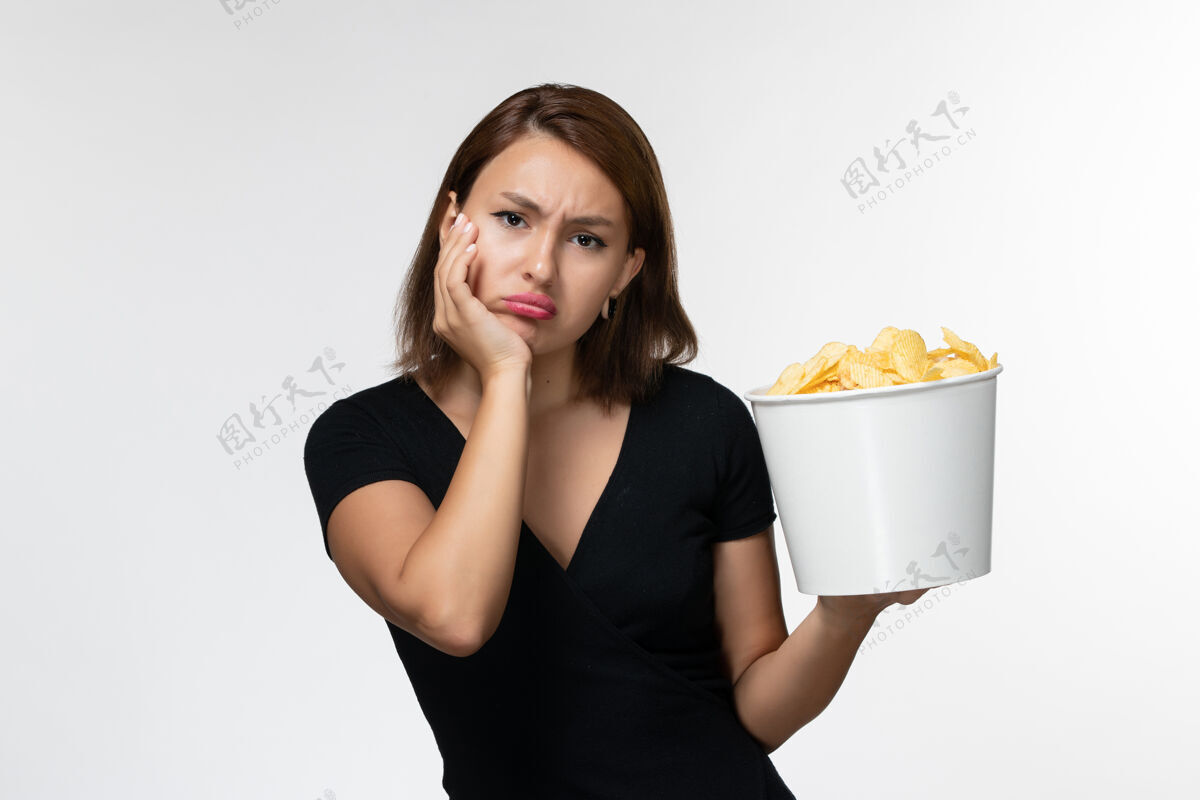 土豆正面图身穿黑色衬衫的年轻女性手持薯片 无聊地站在白色表面上衬衫举行无聊