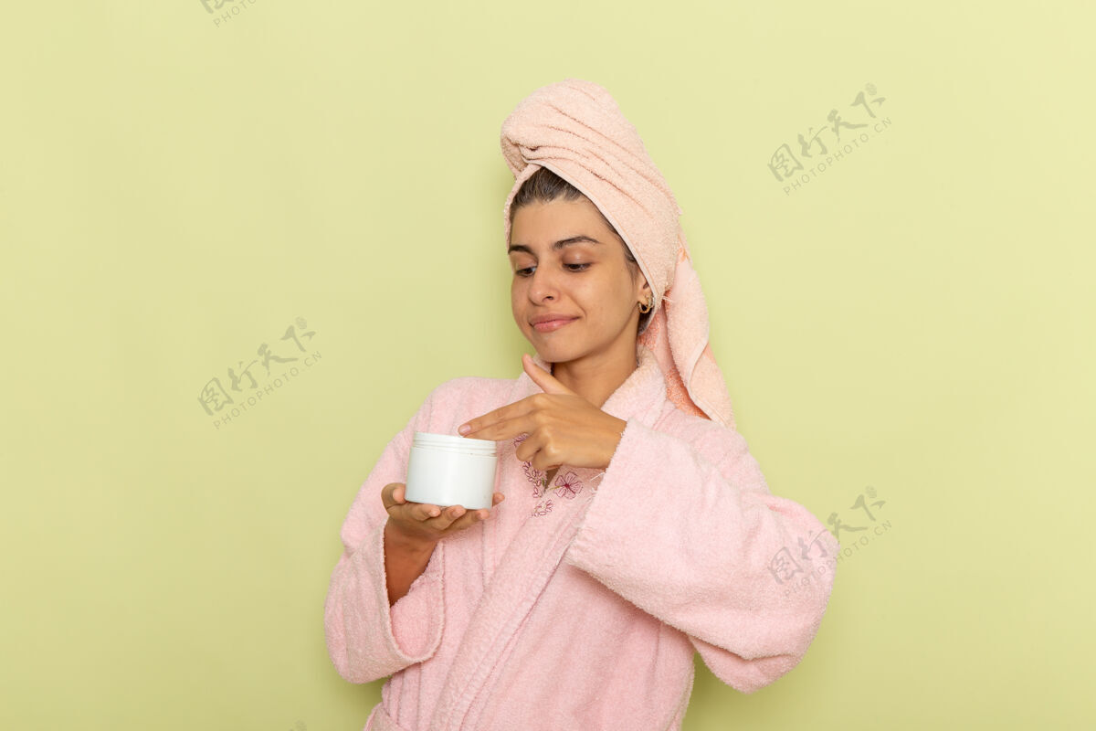 抱着正面图穿着粉红色浴袍的年轻女性 在绿色表面上拿着奶油前面自我照顾浴缸