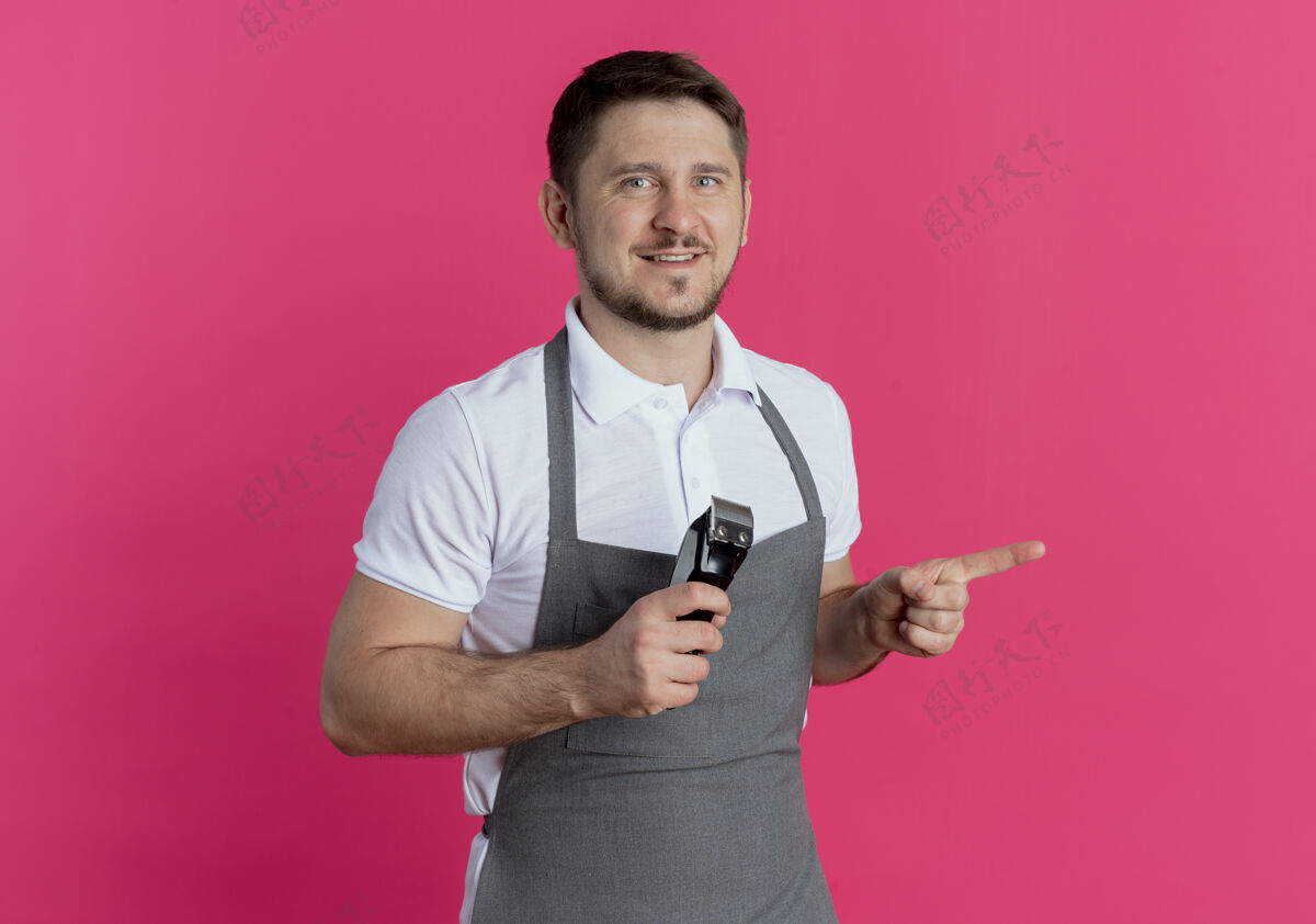 男人围裙上的理发师拿着剃须刀 手指指向一边 微笑着站在粉红色的墙上微笑站着理发师
