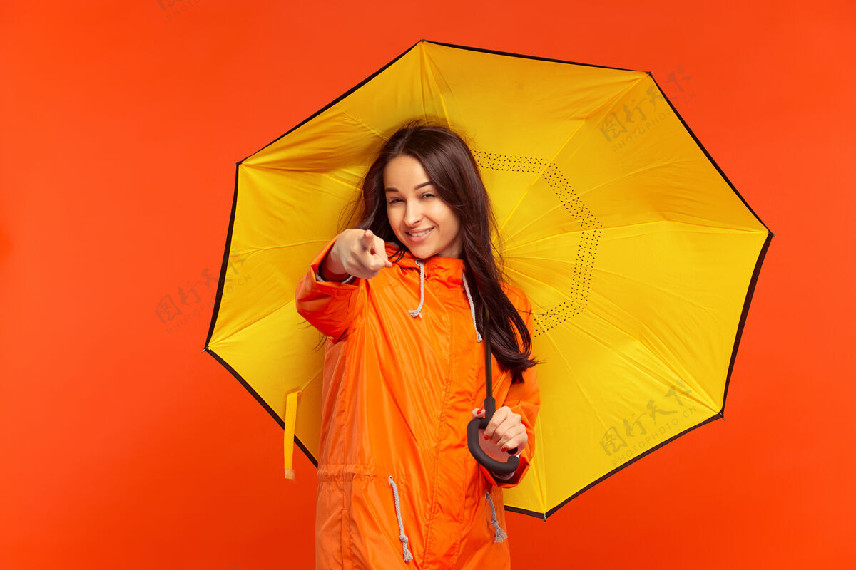 雨那个快乐微笑的小女孩穿着秋天的橘色夹克 指着橘色墙壁上孤立的前面 在摄影棚摆姿势下雨天气快乐