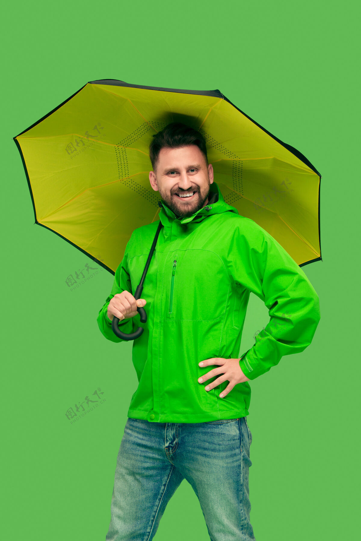 站立帅哥留着胡子 微笑着 拿着雨伞 看着前面孤立的绿色工作室 生动时尚夹克成人时尚